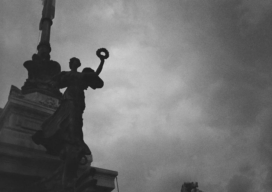 Winged statue silhouetted against heavy clouds, film-like grain