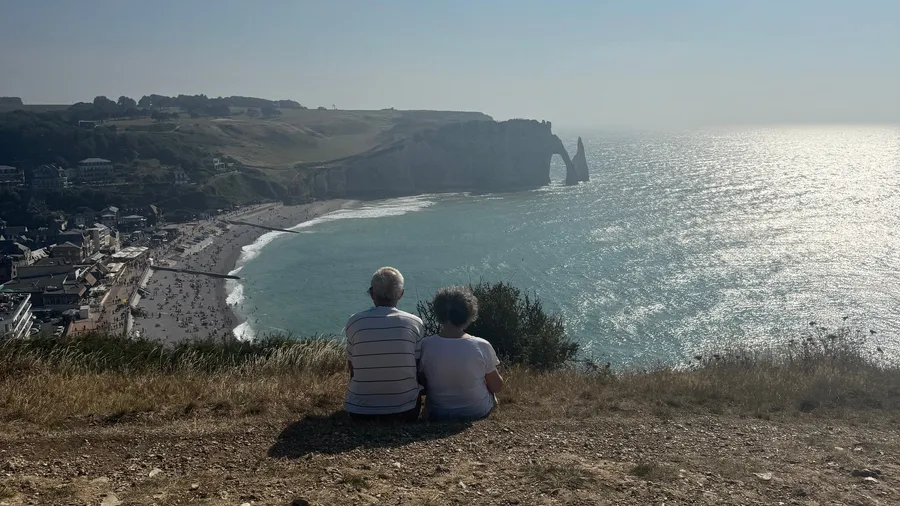Elderly couple seen from behind, sitting together facing the Étretat arch
