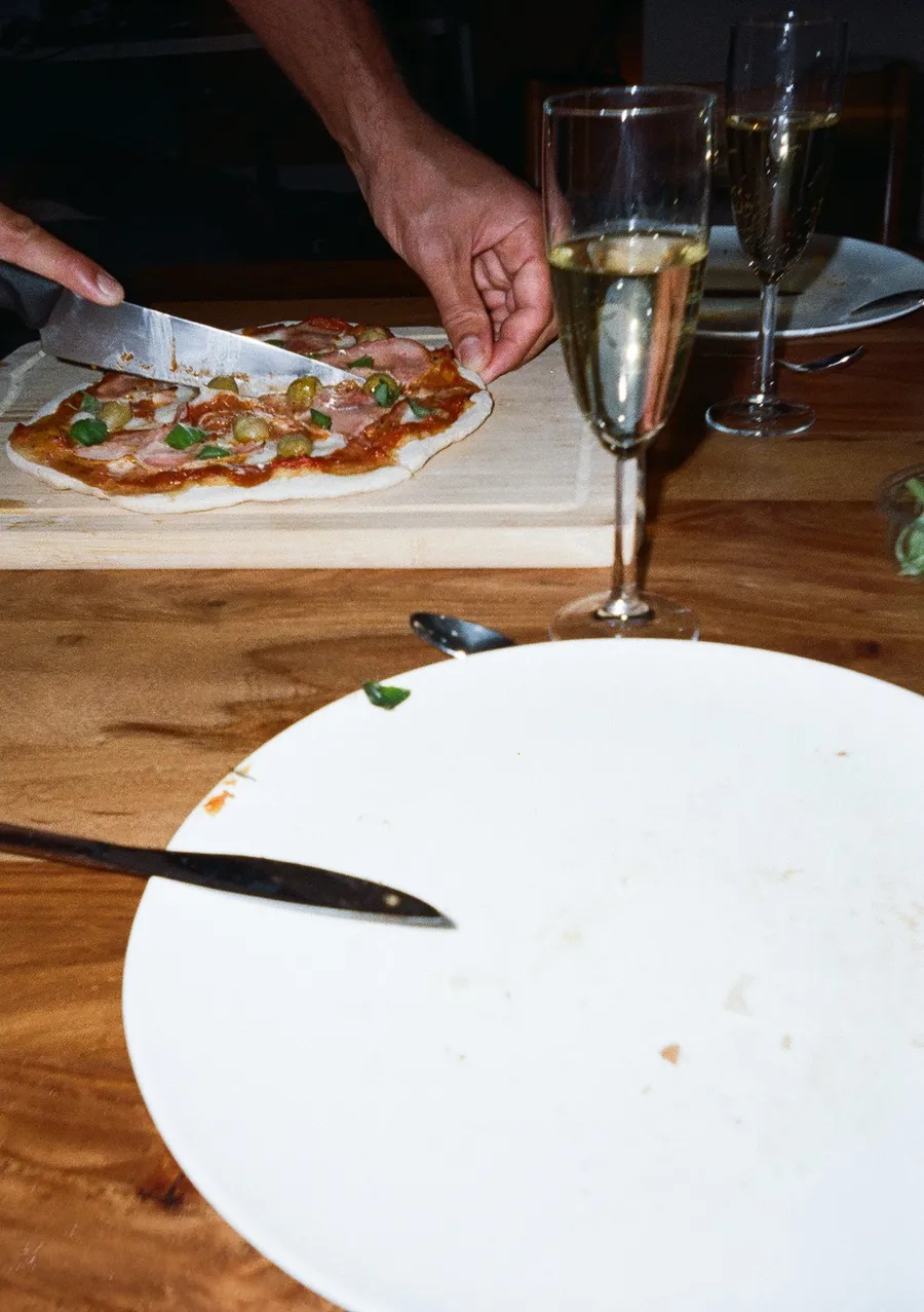 Hands slicing homemade pizza, champagne glasses, empty plate