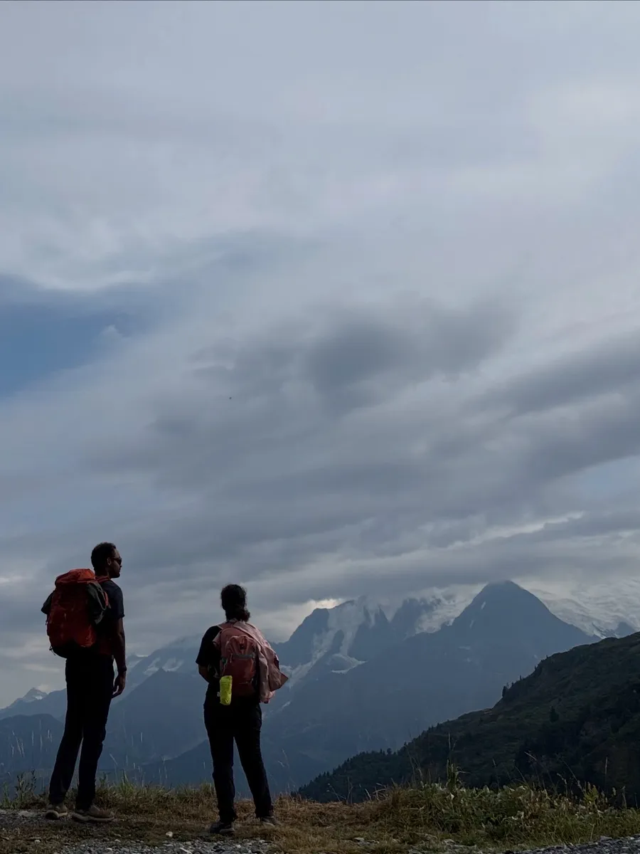 Two hikers facing snowy peaks under grey sky