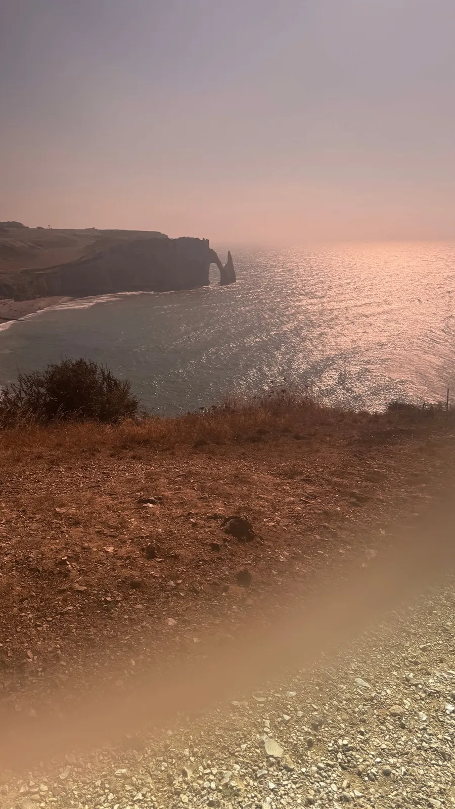 Étretat needle in pink haze, a soft finger-blur glowing in the corner